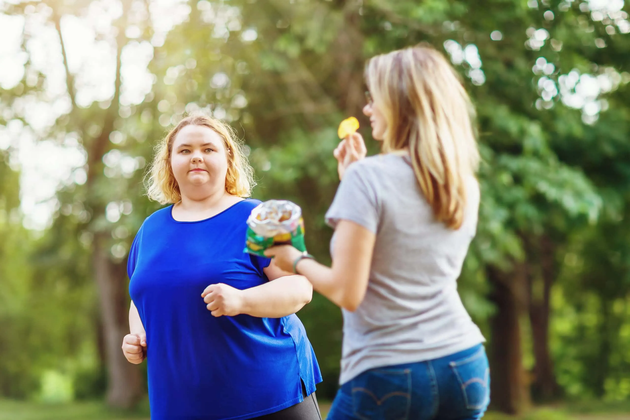 A Young Blonde Of Plus Sizes Runs In The Park Near A Woman Eating Chips. The Concept Of A Healthy Lifestyle And Self Improvement